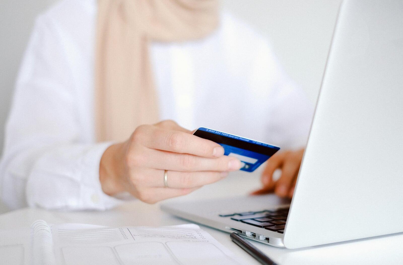 A person with a credit card in their hand, sitting at their laptop ready to make a purchase.